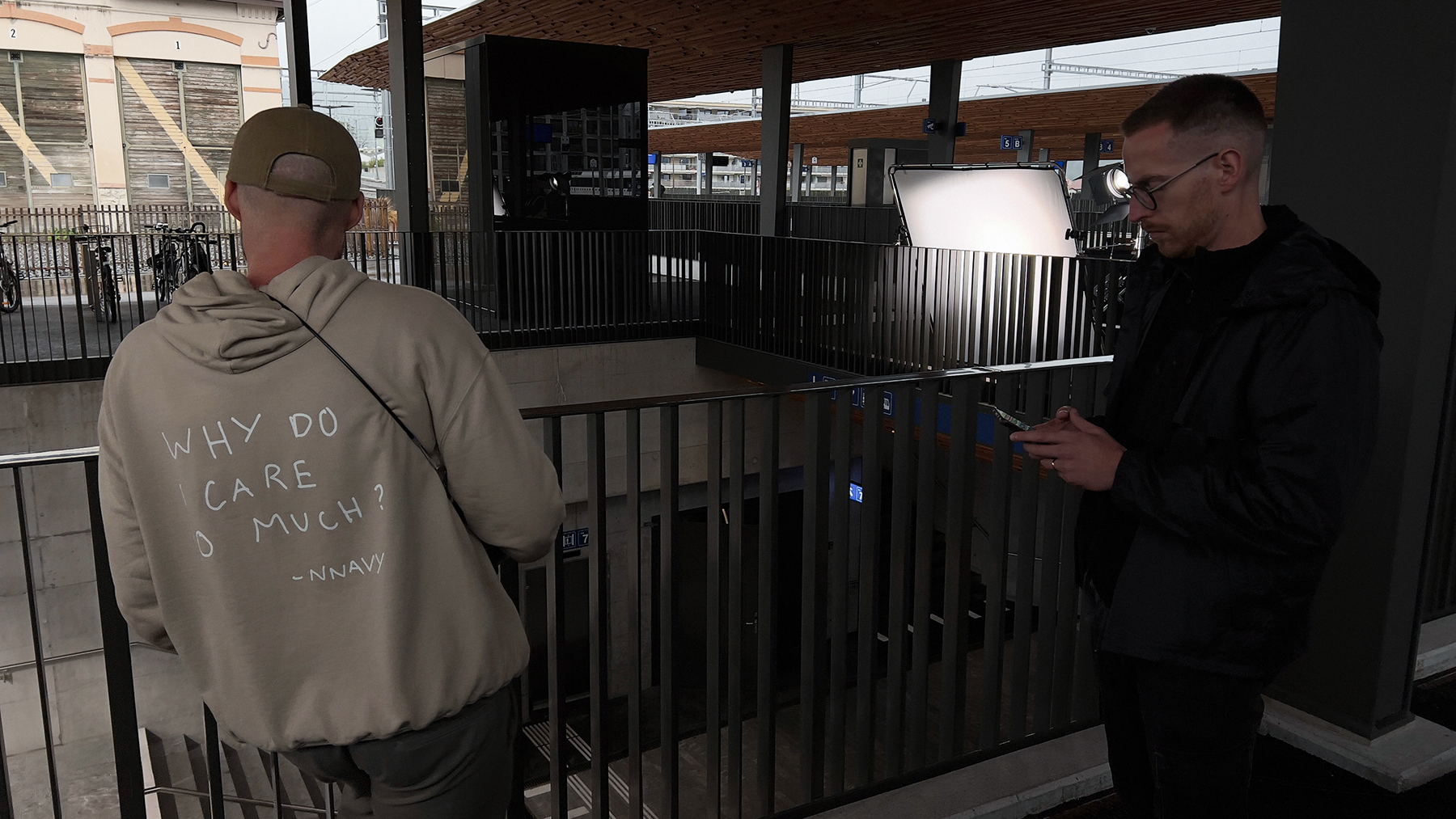 Film crew on a station platform: one man in a khaki hoodie faces away, another checks his phone near a softbox light.
