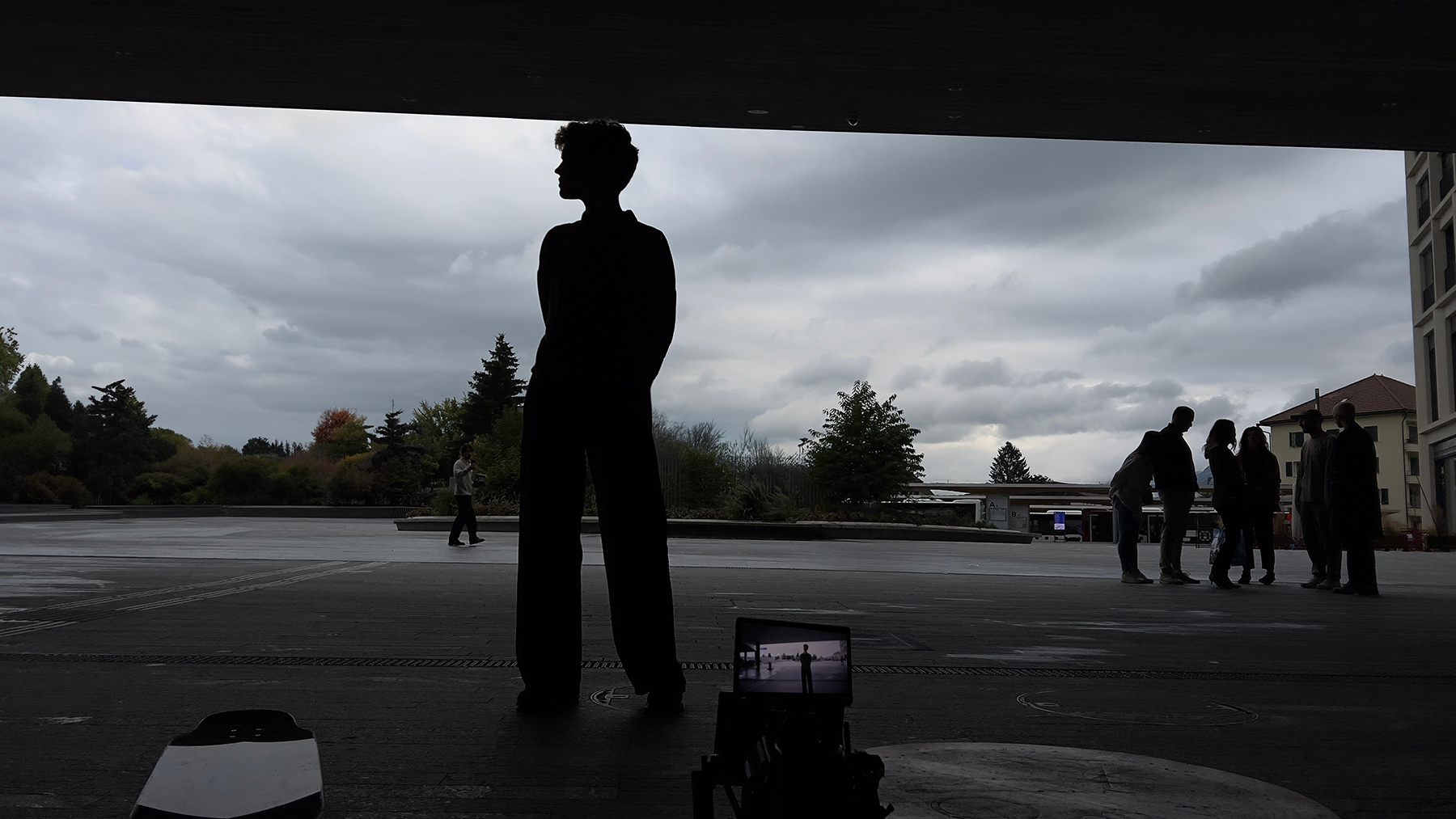 Silhouette of a figure in wide-leg trousers under a shelter, framed on a monitor next to a skateboard as the crew gathers on the square behind.