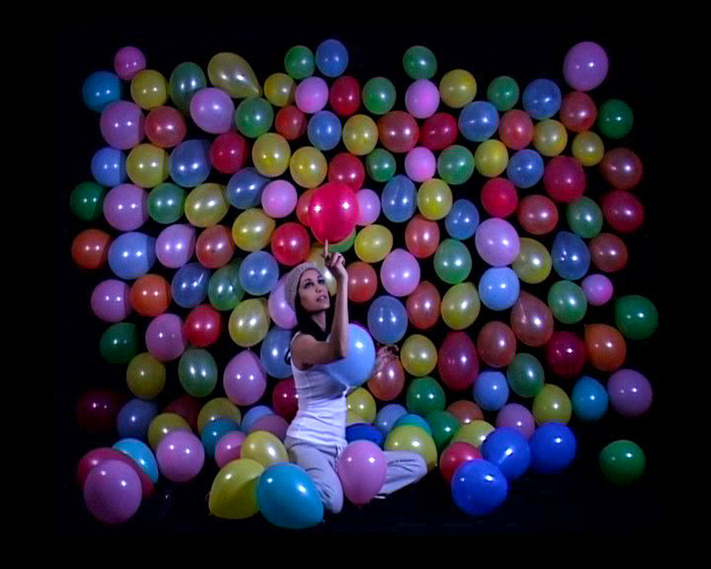 Woman in a beanie kneeling before a wall of colorful balloons, balancing a red balloon on her index finger.