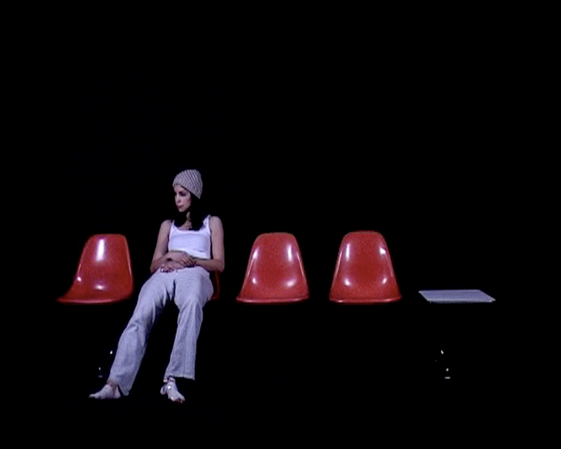 A woman sits alone on the second of four orange bucket chairs in a dark room, arms crossed, legs extended, gazing to the side.